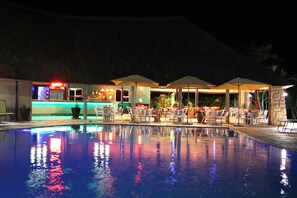 A nighttime pool area with lounge chairs and umbrellas, a bar with neon lights, and a reflection on the pool's surface. - Protea Hotel by Marriott Umfolozi River (Mfolozi)