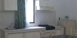 A small kitchen with a window, a sink, cabinets, and a chair.