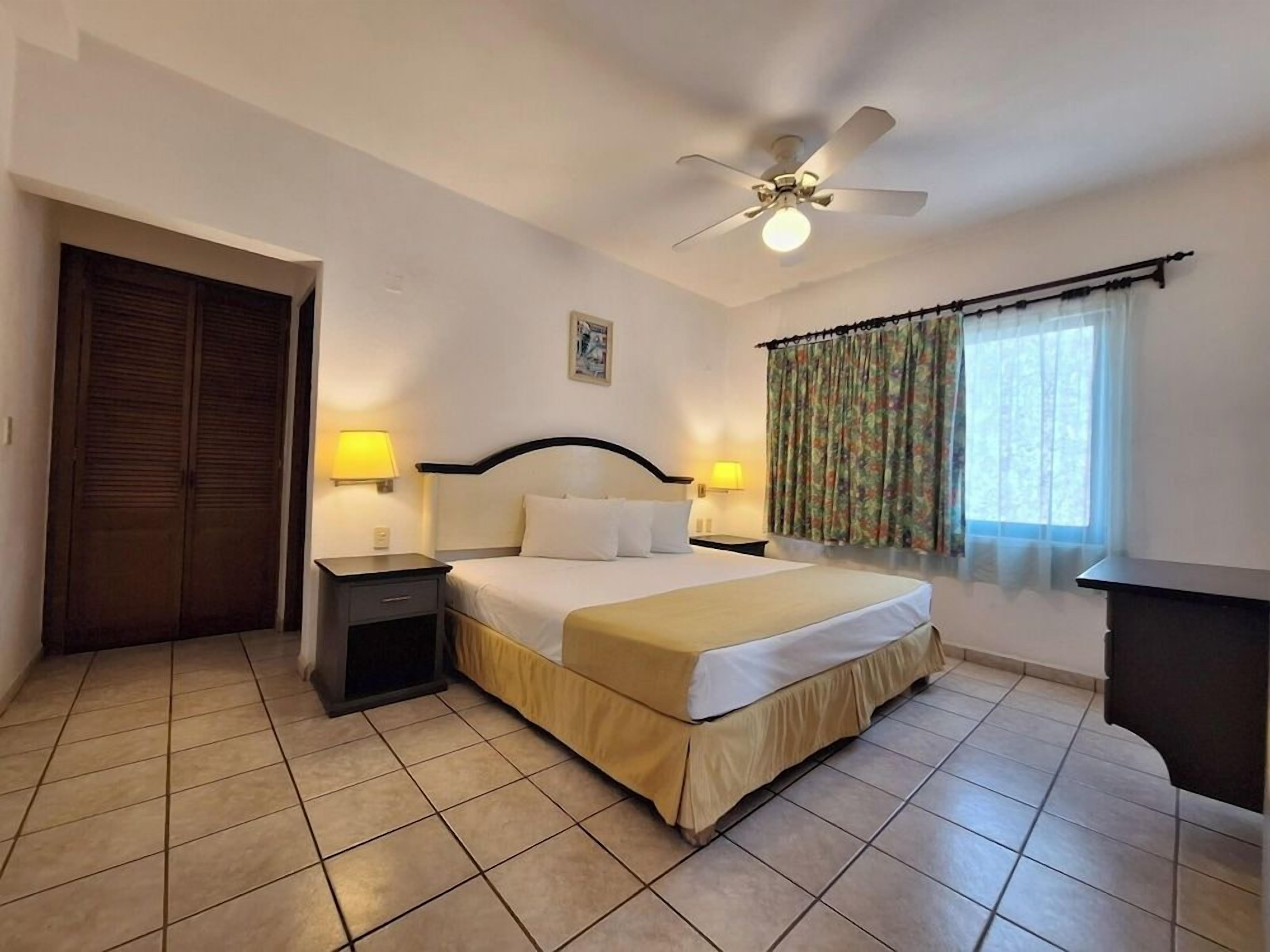 Una habitación de hotel con una cama, dos mesitas de noche con lámparas, un ventilador de techo, una ventana con cortinas y una puerta.