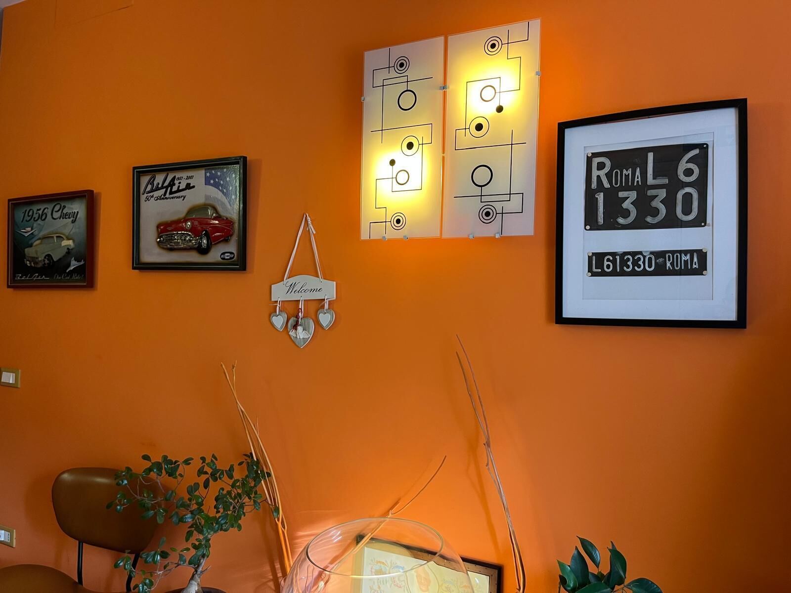 A wall with various framed items including a vintage car poster, a welcome sign, a framed license plate, and abstract wall art.