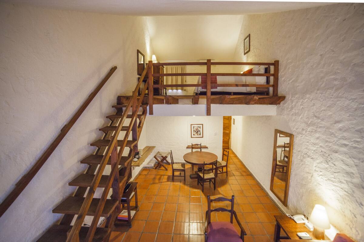 Ein Zimmer mit einer Holztreppe, einem runden Tisch mit Stühlen und einem Bett auf der oberen Ebene.