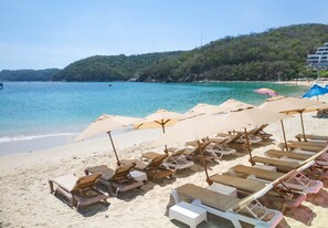 A beach with rows of sun loungers and umbrellas, a clear blue sea, and a forested hill in the background.