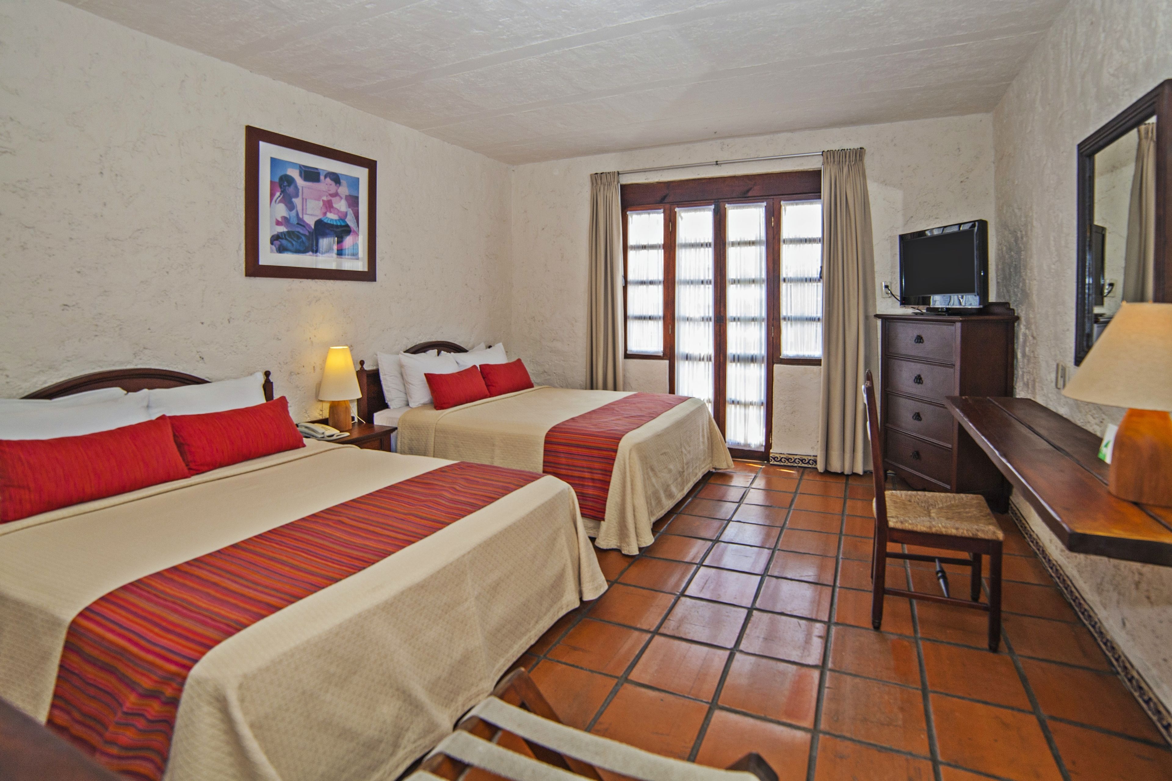 Una habitación de hotel con dos camas, televisión, escritorio con lámpara y ventana con cortinas.