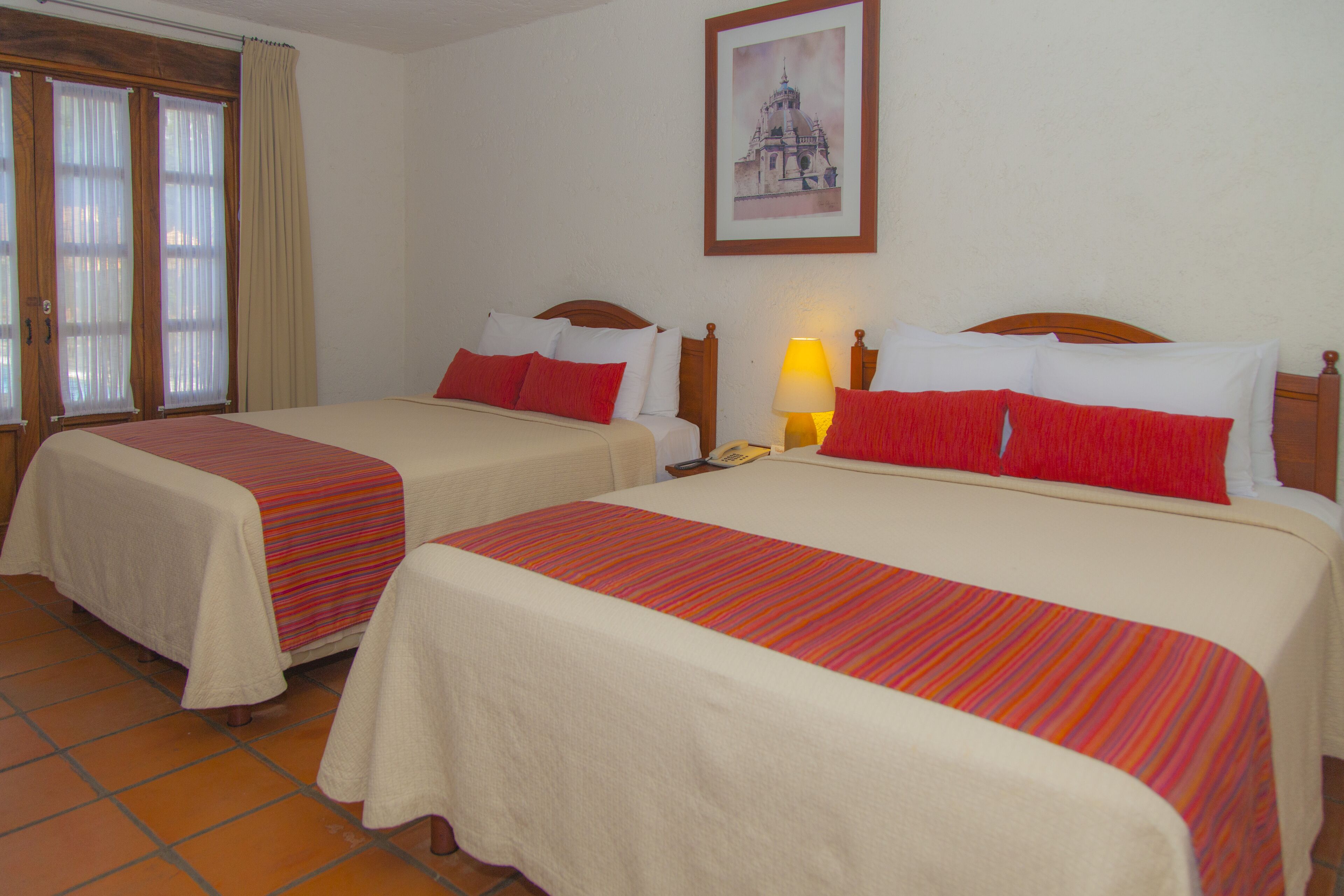 A hotel room with two beds, each with a red and white bedspread, a nightstand with a lamp, and a window with curtains.
