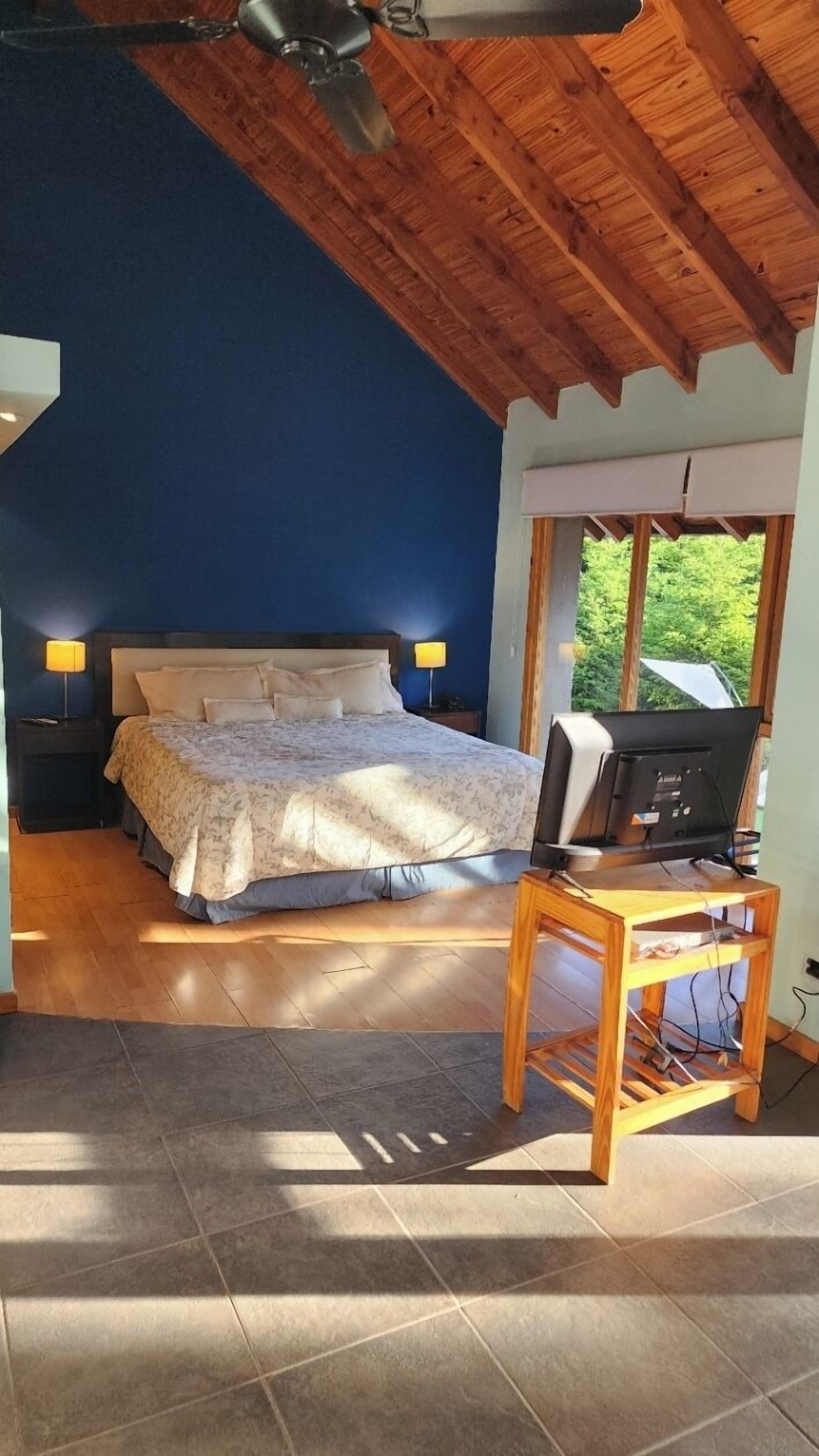 A bedroom with a bed, a desk with a computer, and a window with a view of greenery.