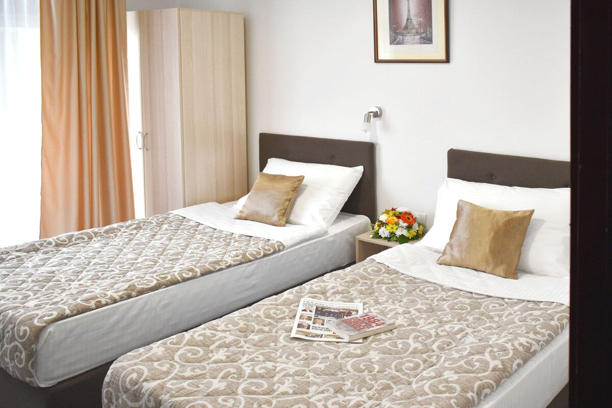 Two single beds with patterned bedding, a nightstand with a vase of flowers, and a framed picture on the wall.