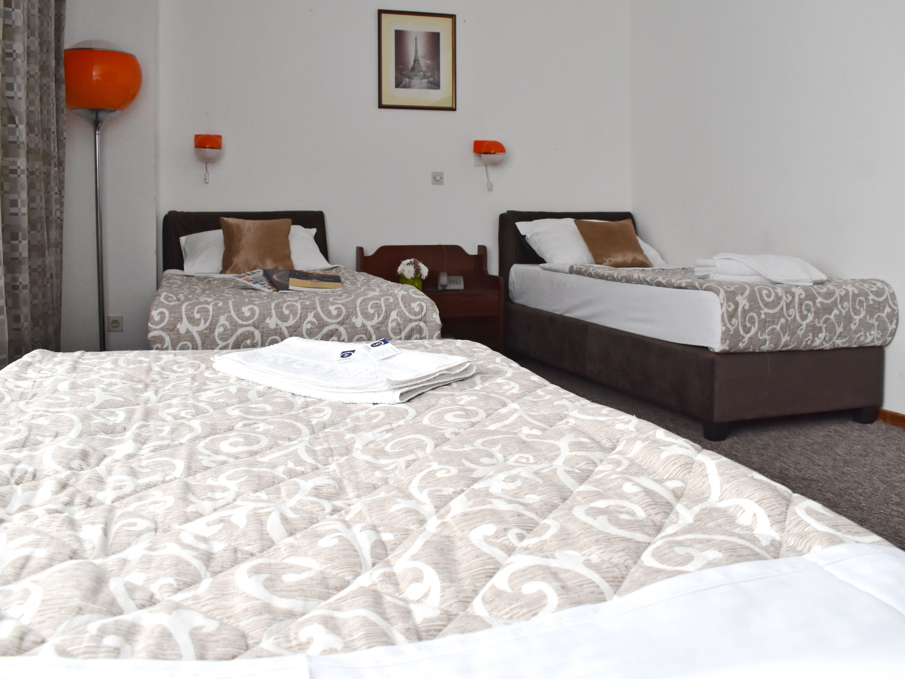 A hotel room with two beds, a large bed in the foreground and a smaller one in the background. There is a nightstand with a lamp between the beds, and a framed picture on the wall above the smaller bed.