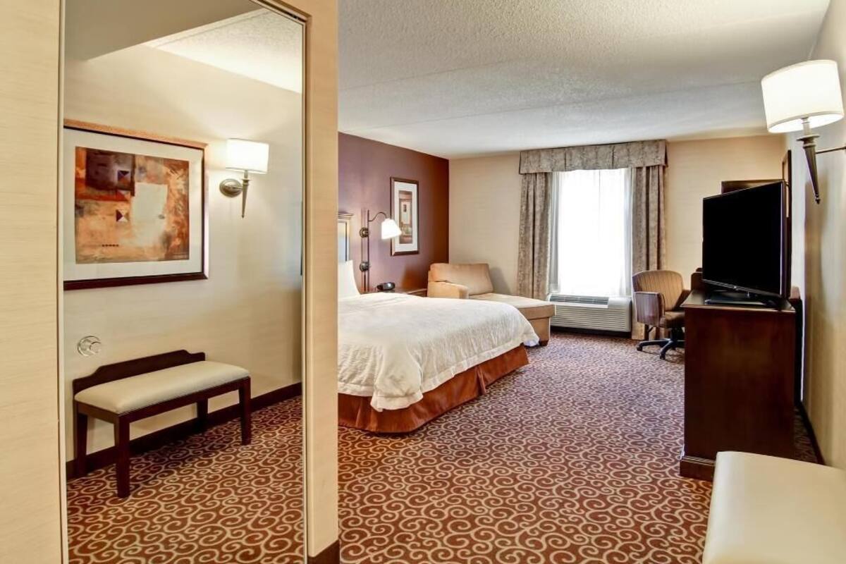 A hotel room with a bed, a desk, a television, and a bench.