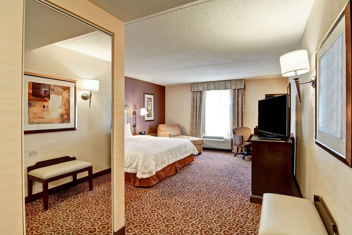 A hotel room with a large bed, a desk with a television, a bench, and a patterned carpet.