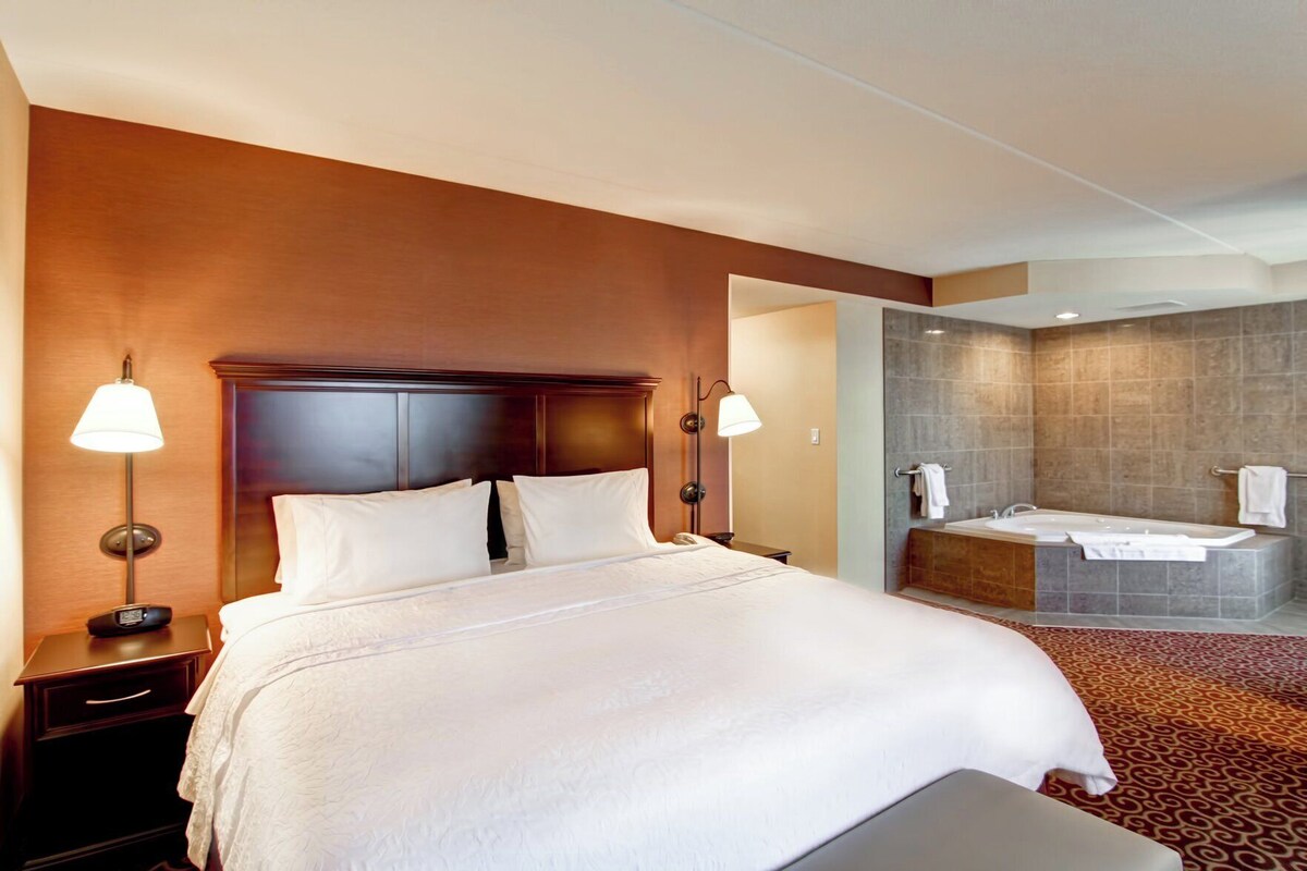 A hotel room with a large bed, bedside lamps, a nightstand, a bathroom with a bathtub, and a shower area.