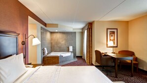 A hotel room with a large bed, a desk, a chair, a desk lamp, a telephone, a bathroom with a bathtub and a shower, and a patterned carpet. - Hampton Inn by Hilton Sudbury (Sudbury)
