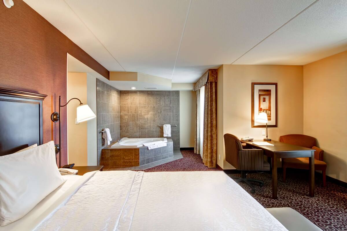 A hotel room with a large bed, a desk, a chair, a desk lamp, a telephone, a bathroom with a bathtub and a shower, and a patterned carpet.