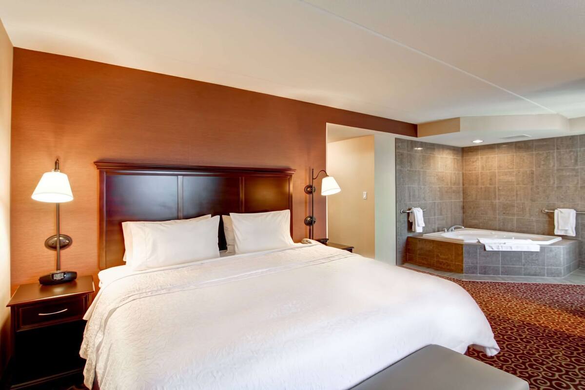 A hotel room with a large bed, bedside lamps, a nightstand, a bathroom with a bathtub, and a shower area.