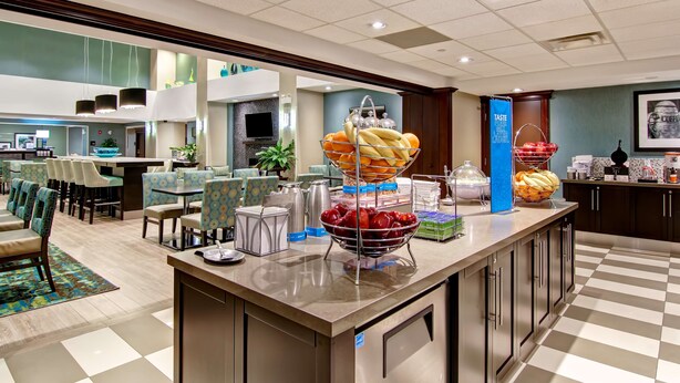 A hotel lobby with a buffet area featuring fruit and bread. There are seating areas with patterned chairs and a checkered floor. - Hampton Inn by Hilton Sudbury (Sudbury)