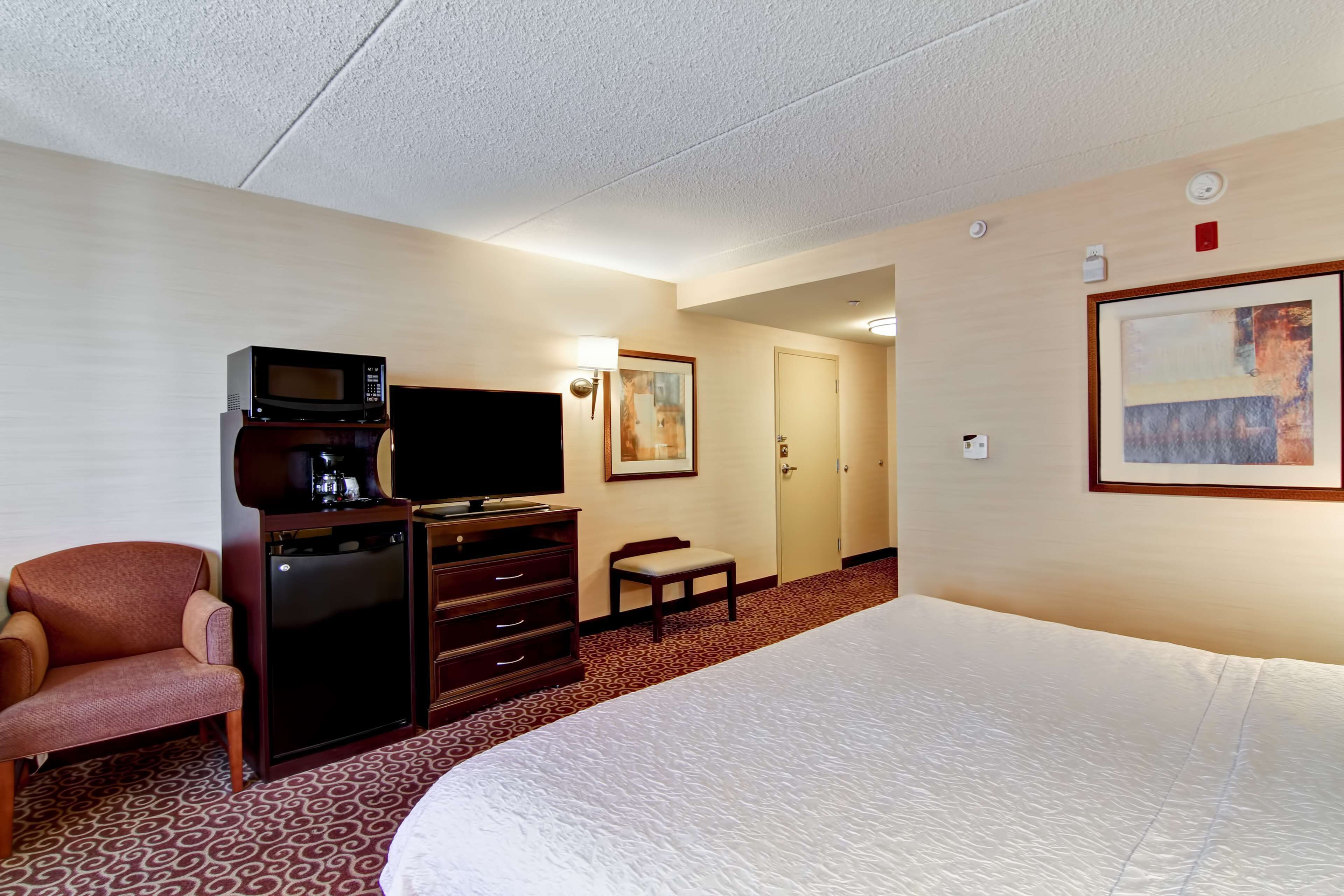 A hotel room with a bed, a television, a coffee maker, a desk with drawers, a chair, a bench, and a door.