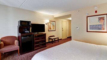 A hotel room with a bed, a television, a coffee maker, a desk with drawers, a chair, a bench, and a door.