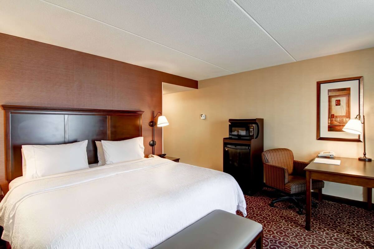 A hotel room with a large bed, a desk with a lamp, a chair, a microwave, and a framed picture on the wall.
