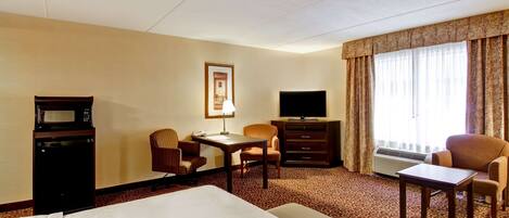 A hotel room with a large bed, a desk with a chair, a television, a microwave, and a window with curtains.