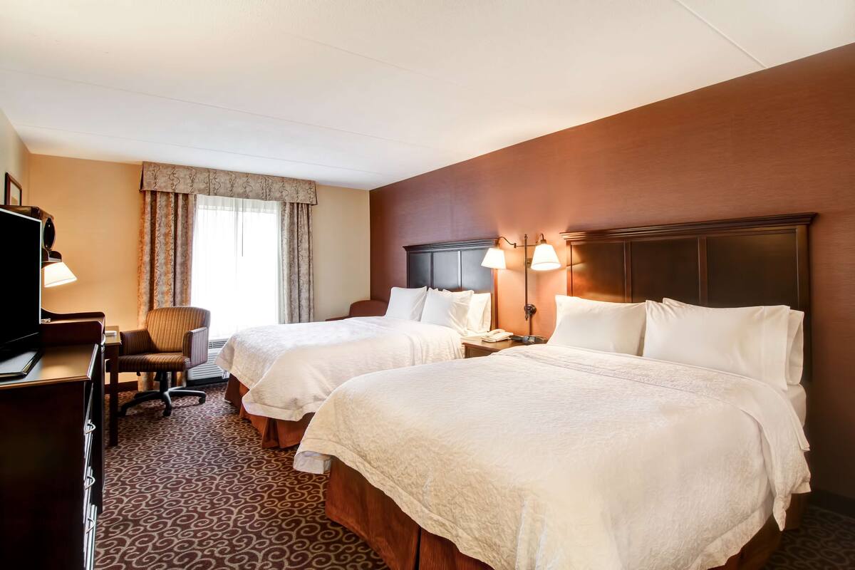 A hotel room with two beds, a desk with a TV, a chair, and a window with curtains.