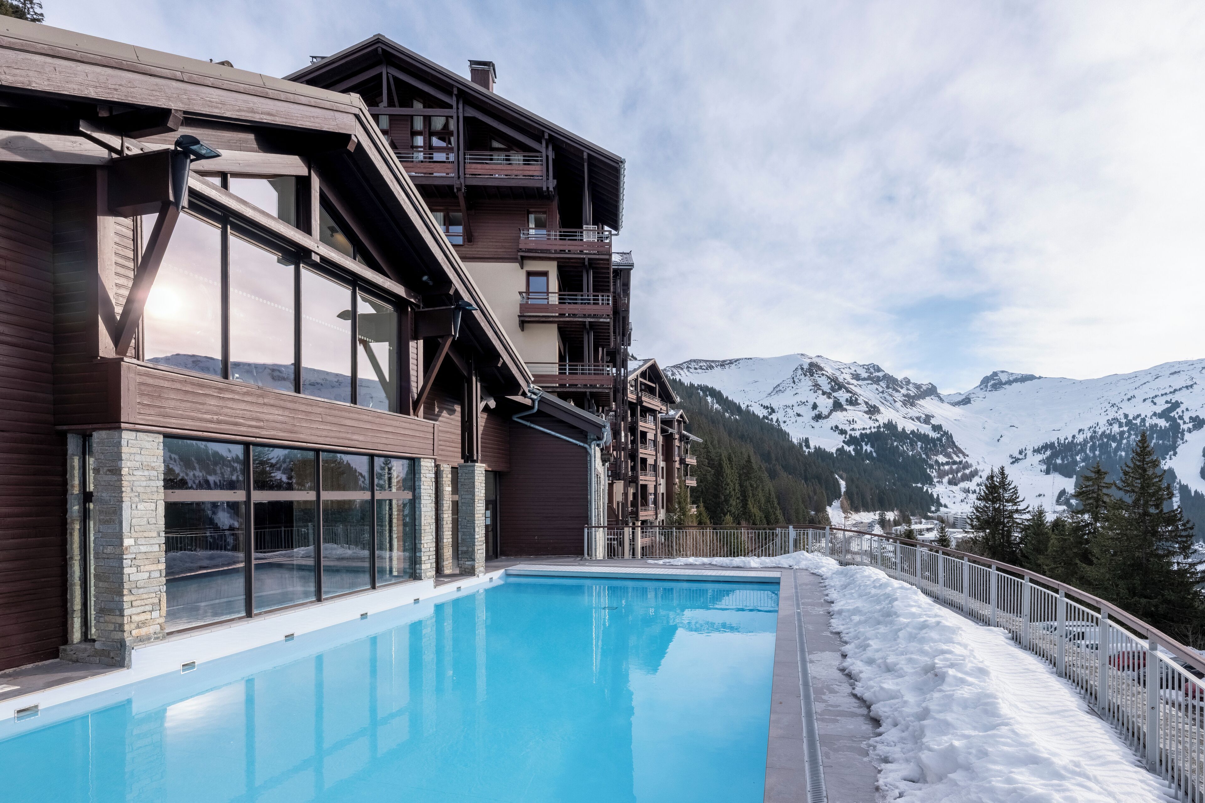 Um edifício moderno com piscina, varandas de madeira e vista para montanhas cobertas de neve.