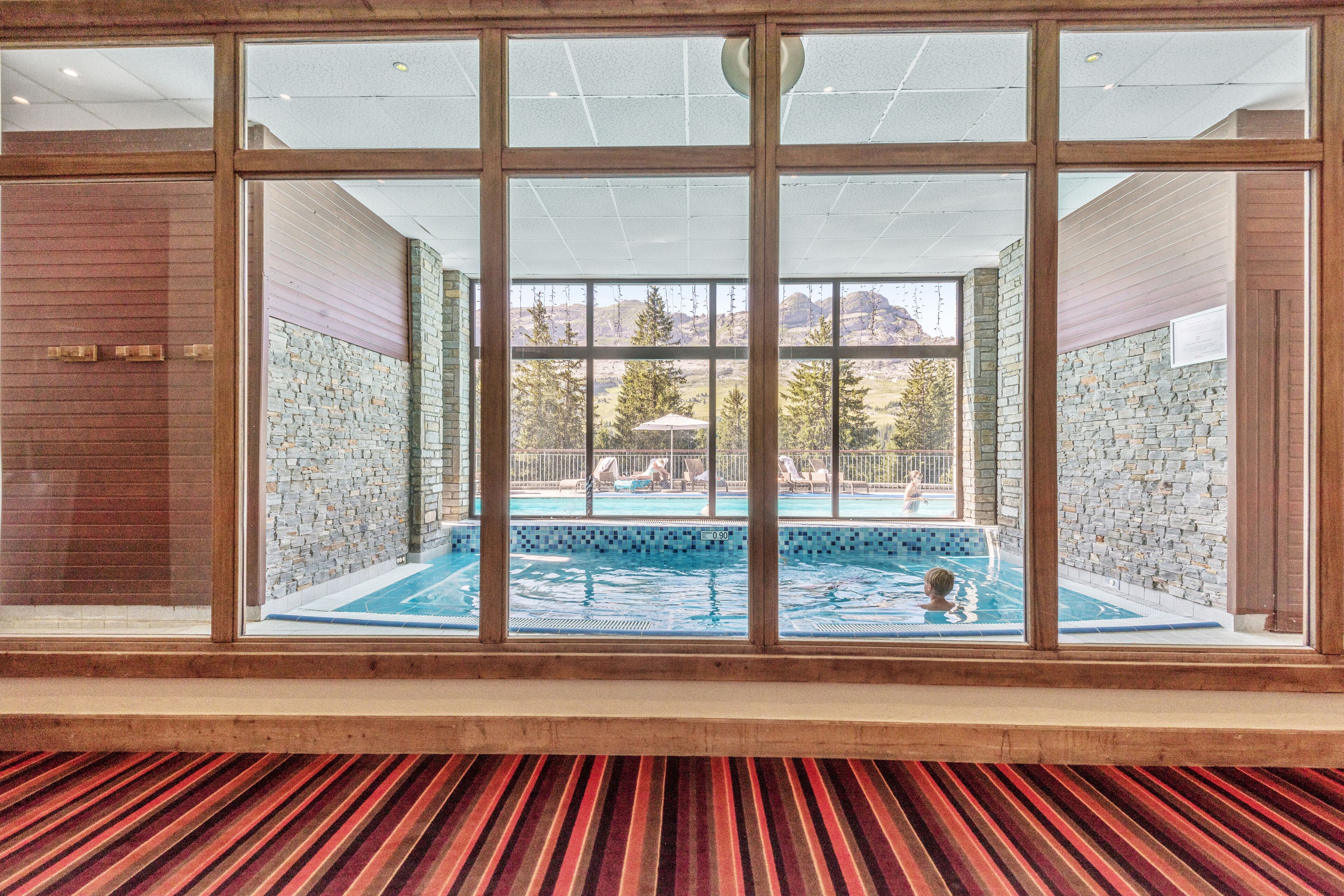A view through large windows of a swimming pool area with stone walls and a view of the outdoors.