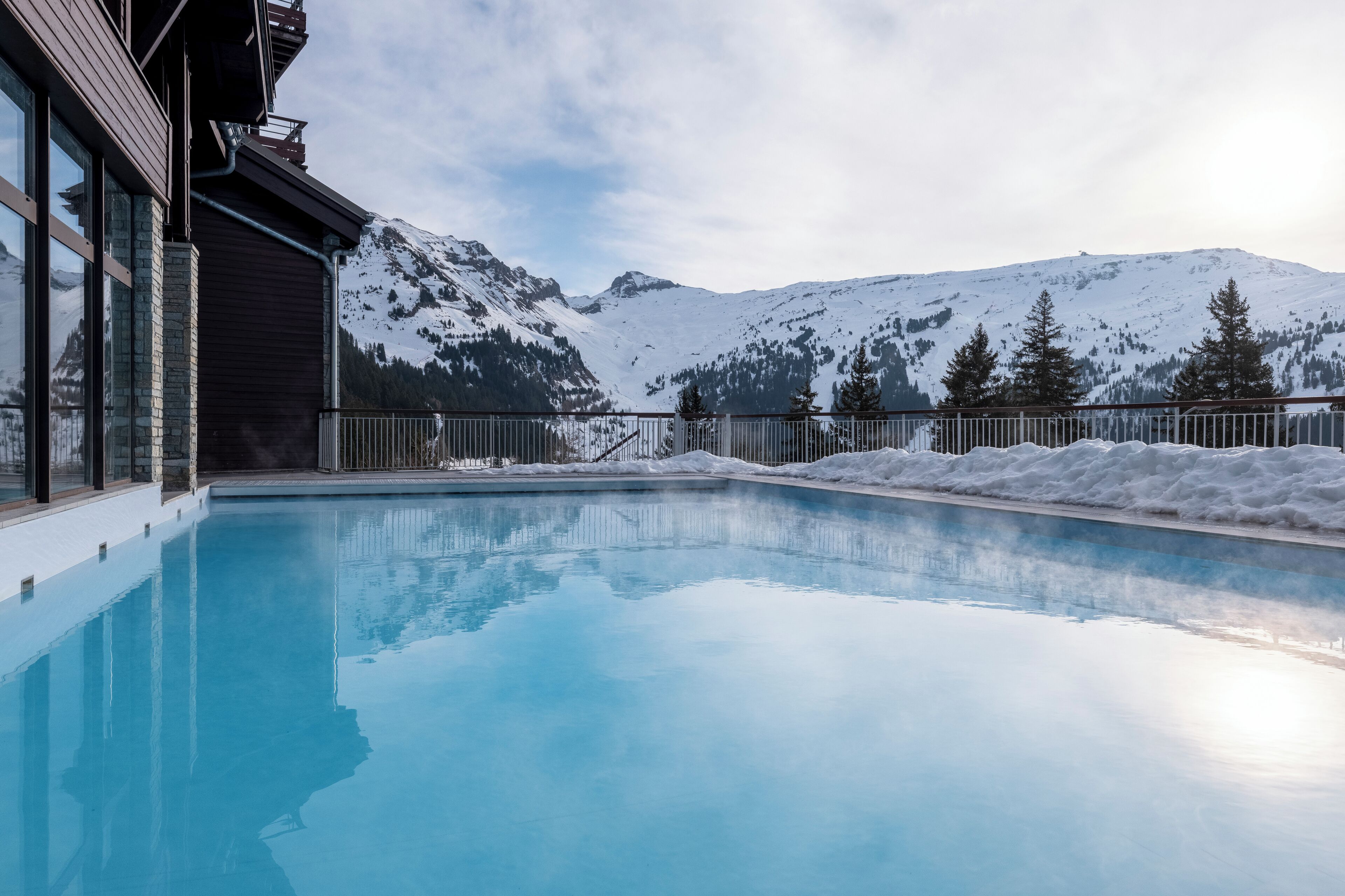 A swimming pool with a mountain backdrop, surrounded by snow and trees.
