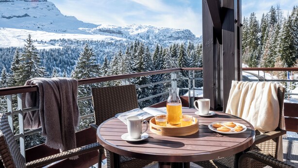 A round wooden table with a tray holding a pitcher, glasses, and cookies, set on a balcony with a view of snow-covered mountains and trees. - Pierre & Vacances Premium Flaine Les Terrasses d'Eos (Araches-la-Frasse)