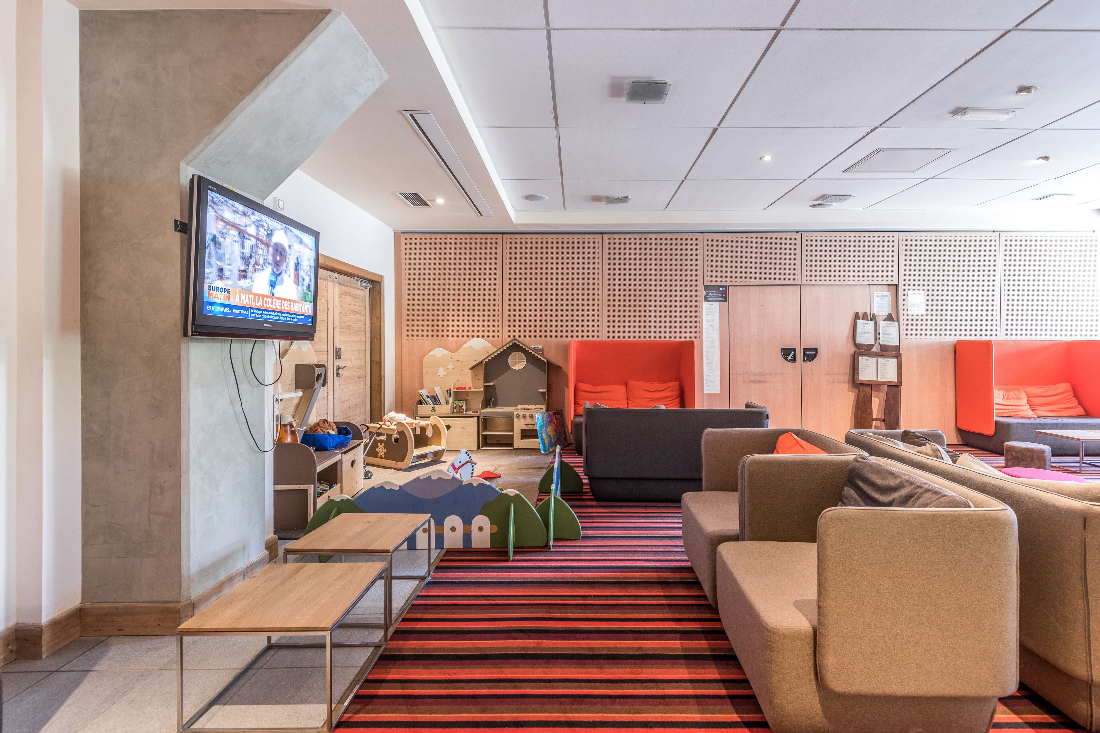 Un rebedor d'hotel modern amb un televisor de pantalla plana, seients confortables, una zona de jocs i una catifa amb ratlles.
