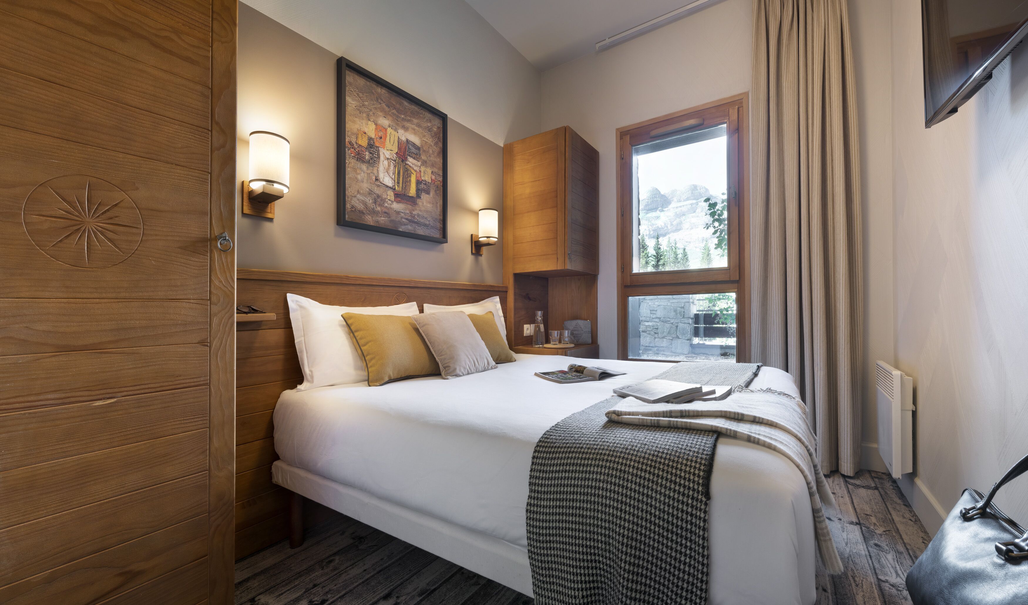 Hotelkamer met een houten hoofdeinde, een groot bed met witte lakens, twee wandlampen, een schilderij boven het bed en een raam met uitzicht op bomen.
