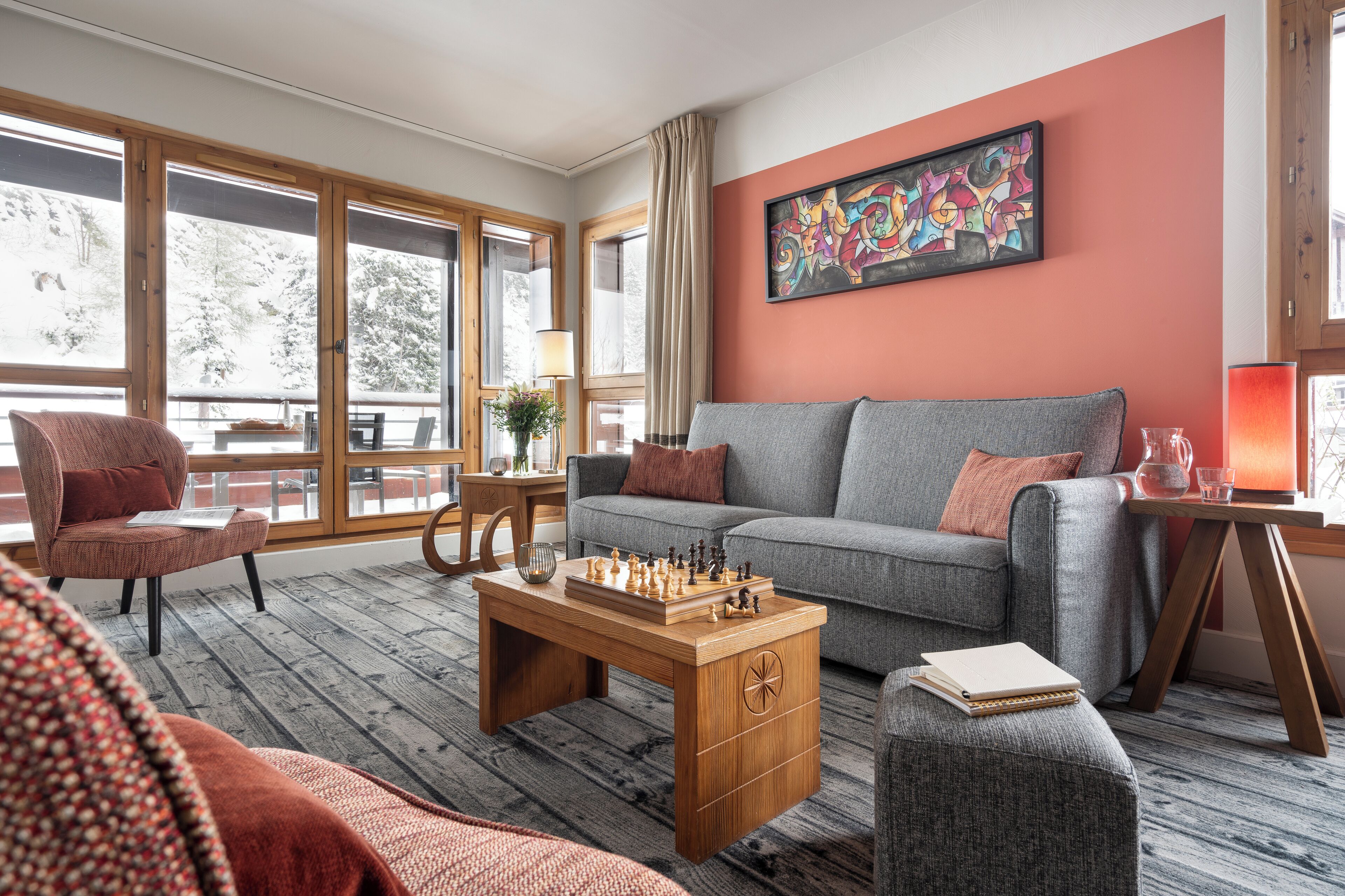 A cozy living room with a grey sofa, a wooden coffee table with chess pieces, and a large window with curtains.