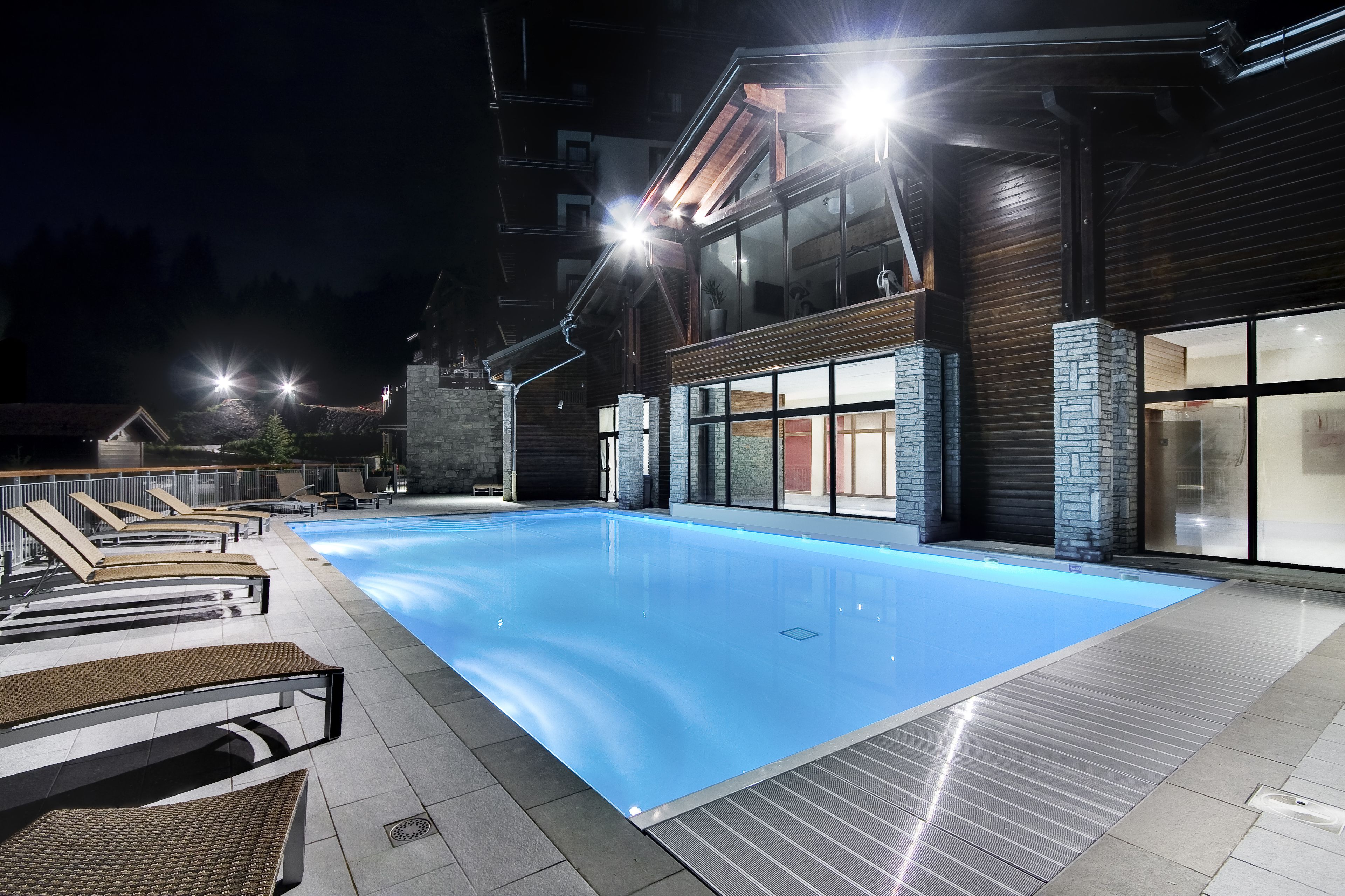A swimming pool with lounge chairs, a stone wall, and a modern building with large windows and a wooden facade.