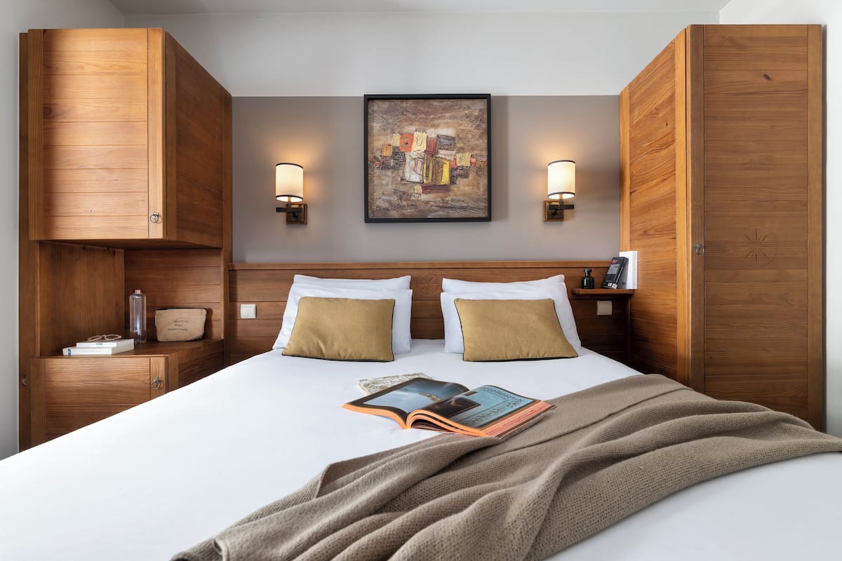 Ein ordentlich bezogenes Bett mit einem Buch, zwei Kissen und einer Decke. Holzschränke und eine Wandlampe sind zu sehen.