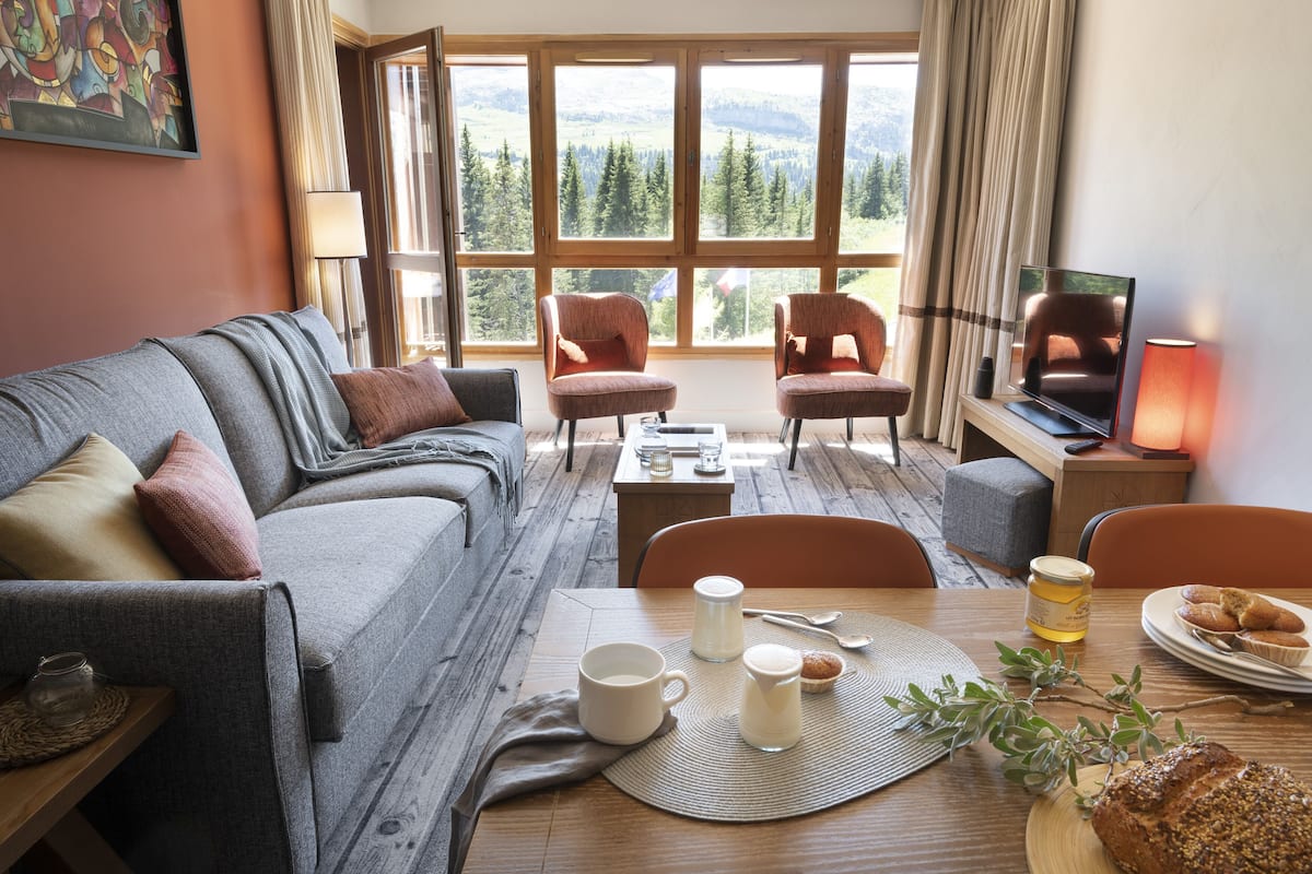 Ein gemütliches Wohnzimmer mit einer Couch, Sesseln, einem Couchtisch und einem Fernseher. Ein Fenster bietet Blick auf Bäume und Berge.