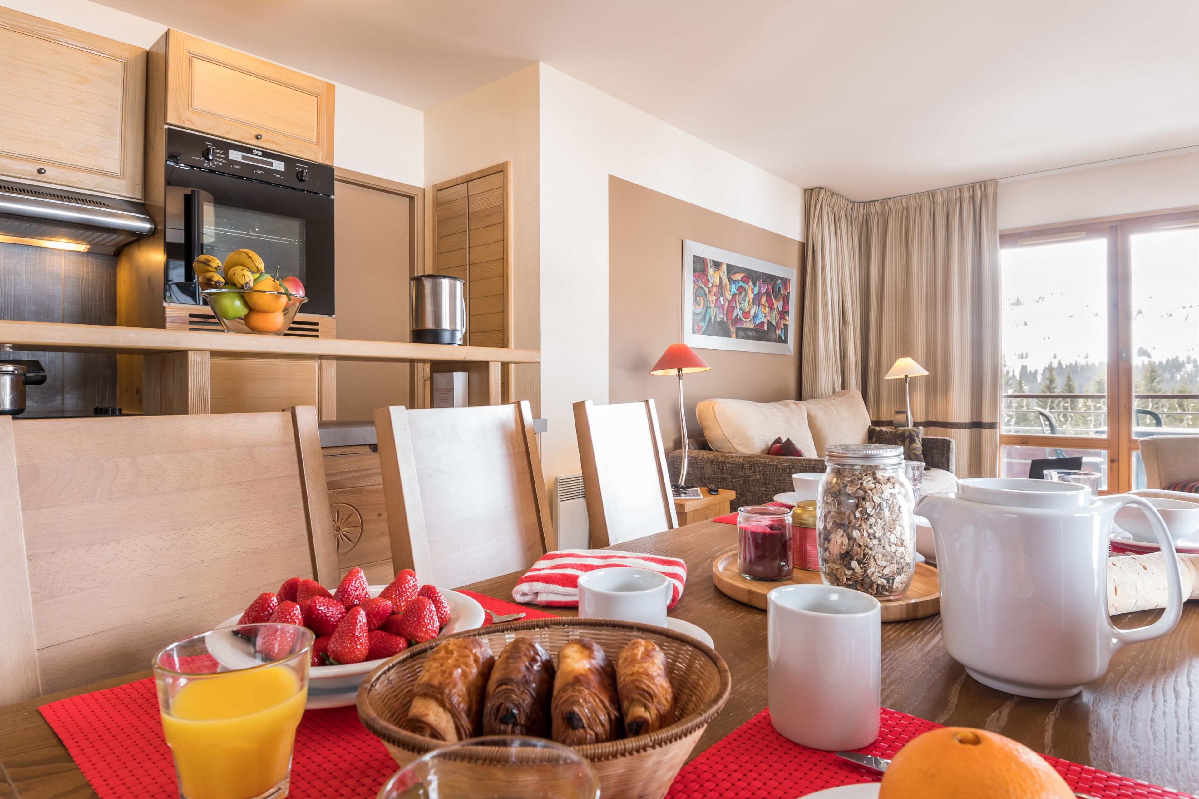 A breakfast setup with fruit, pastries, and coffee in a modern kitchen and dining area with a view.