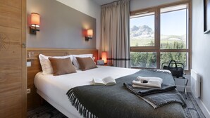 A neatly made bed with a book, pillows, and a blanket. There are two wall-mounted lamps, a window with curtains, and a view of mountains. - Pierre & Vacances Premium Flaine Les Terrasses d'Eos (Araches-la-Frasse)