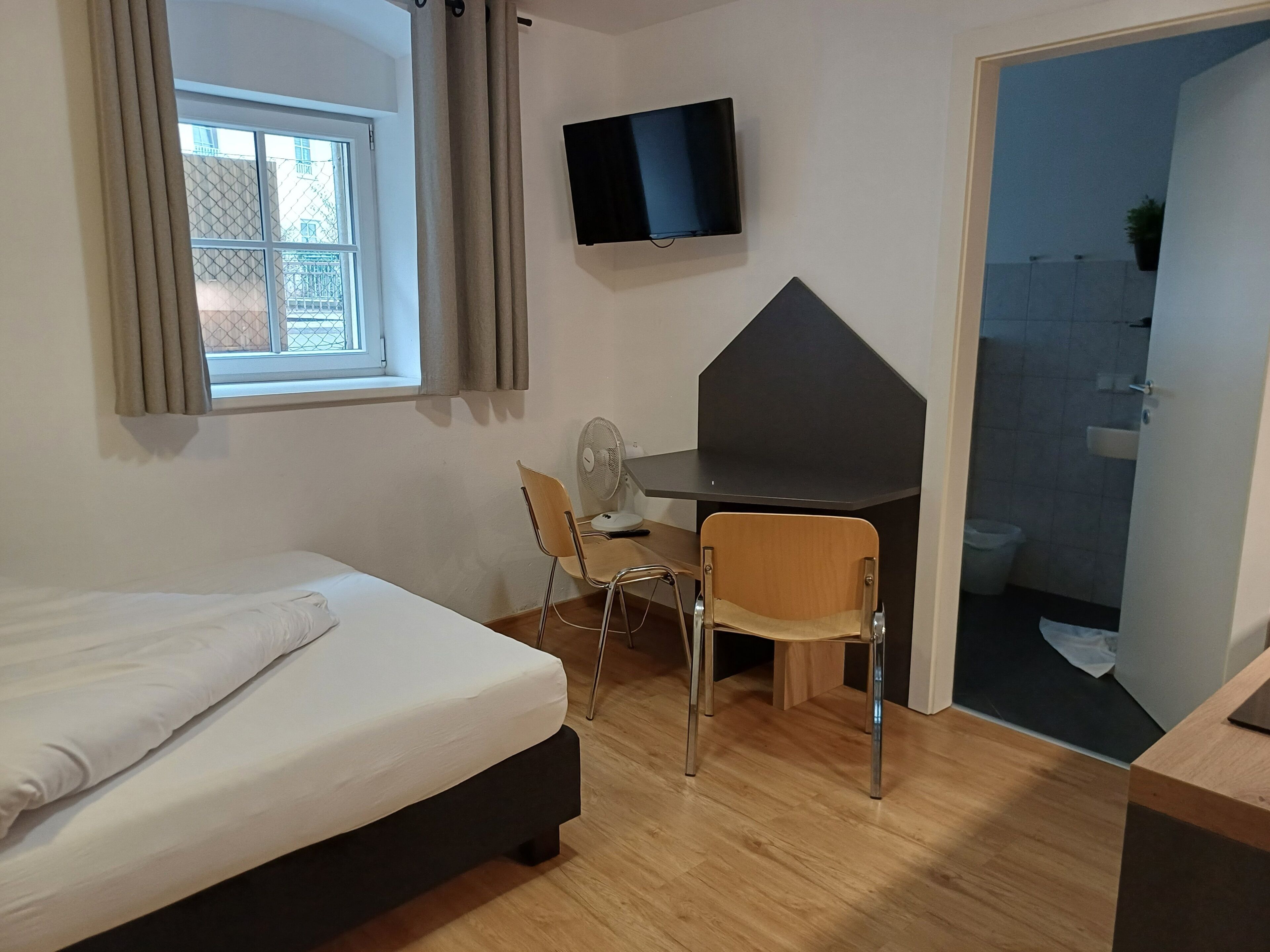 Quarto com cama, escrivaninha, duas cadeiras, televisão e banheiro visível por uma porta aberta.