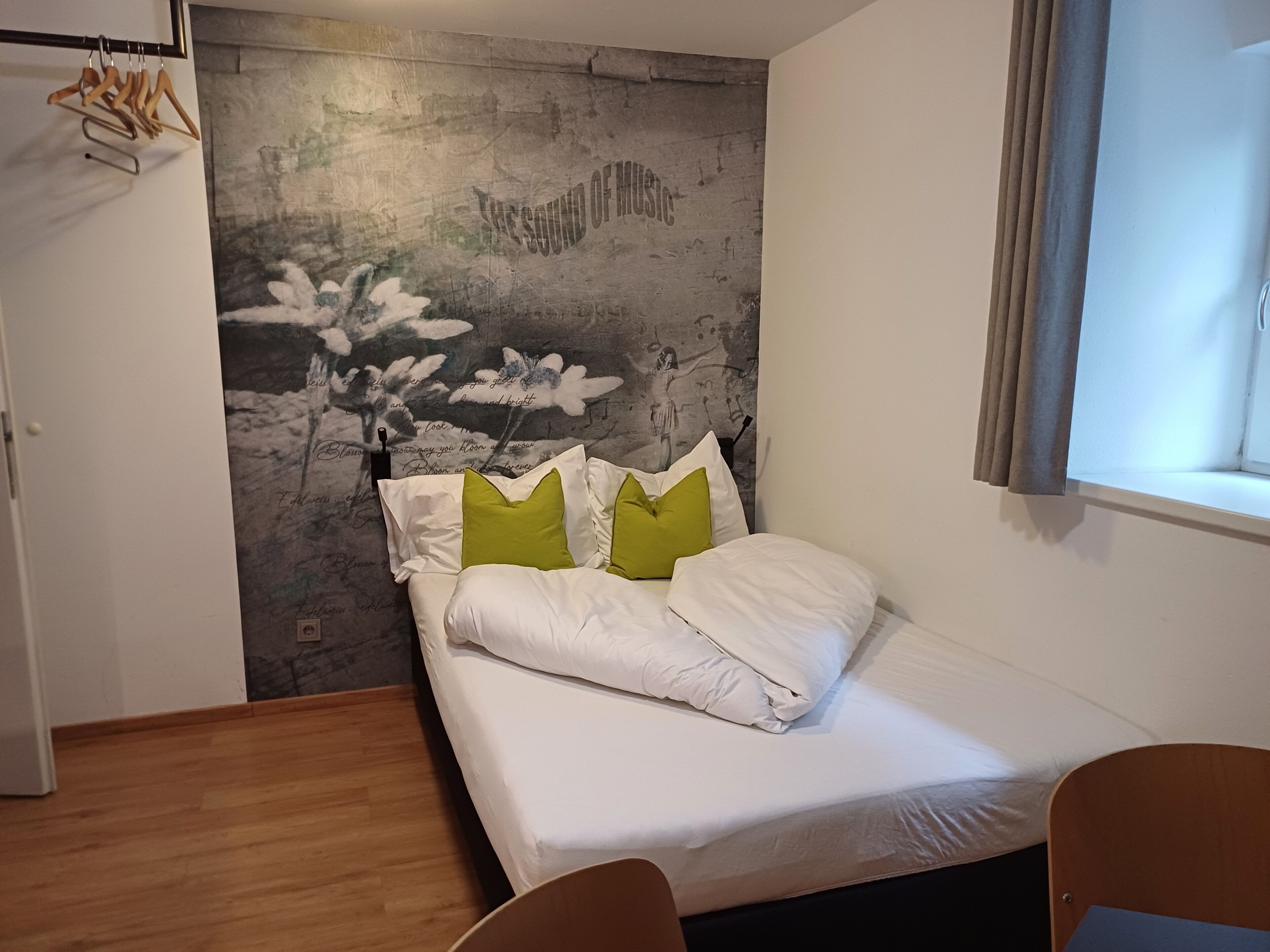 Ein Schlafzimmer mit einem Bett, Holzboden, Wand mit floralem Wandbild und einem Fenster mit Vorhängen.