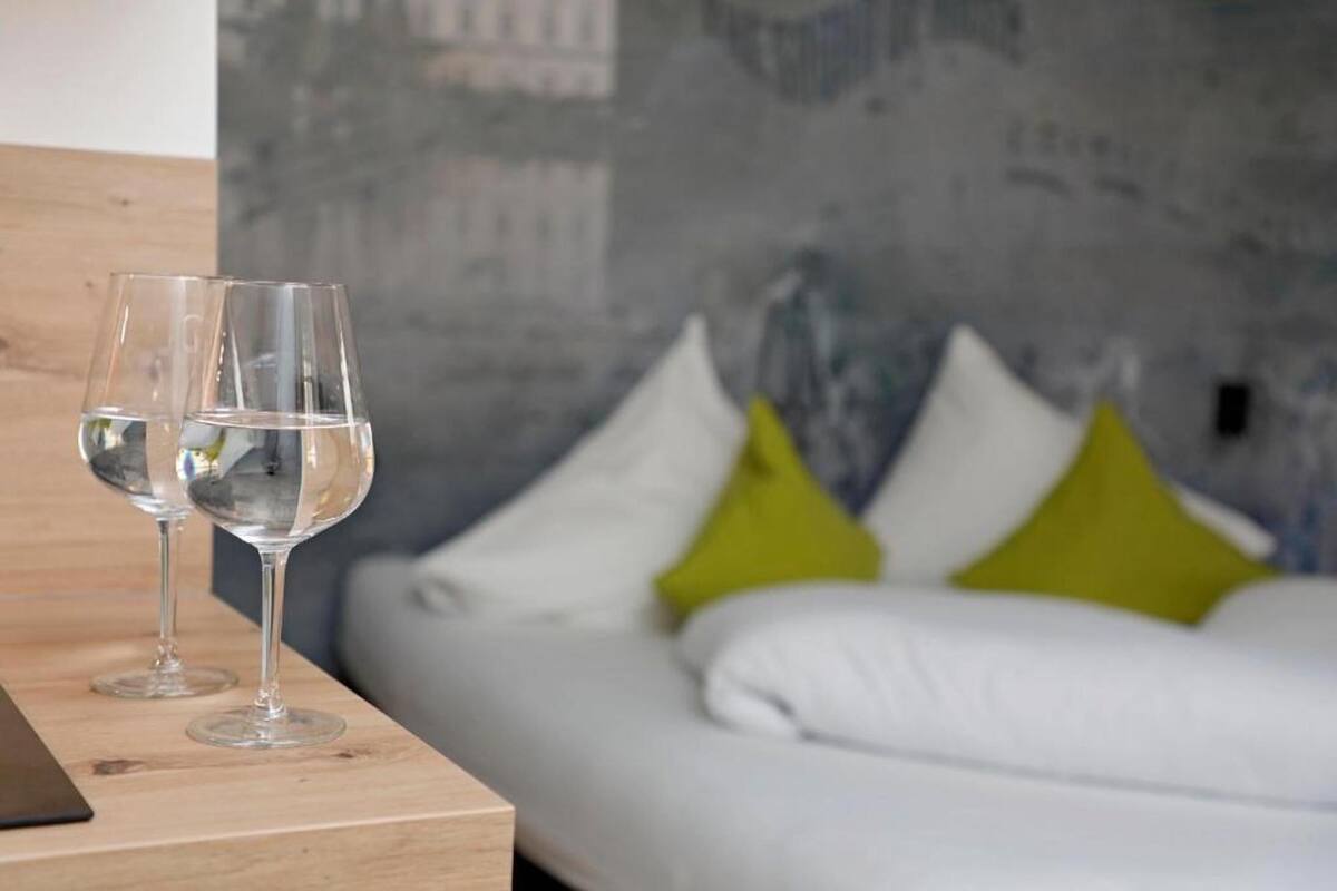 Une pièce avec un lit, deux verres à vin vides posés sur une table en bois et un mur gris à la texture particulière.