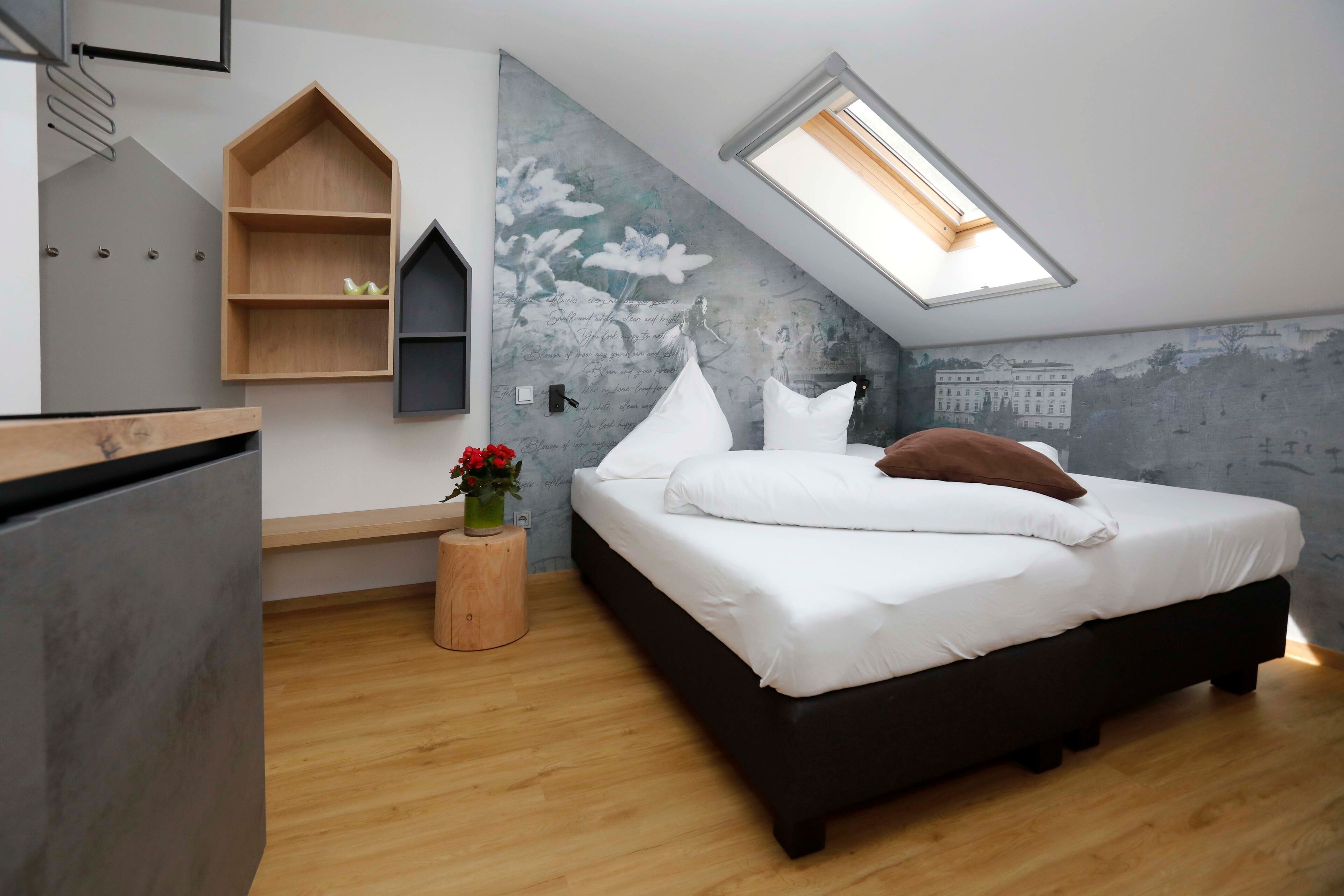 A modern bedroom with a sloped ceiling, a skylight, a bed with white linens, a wooden nightstand, and a wall-mounted shelf.