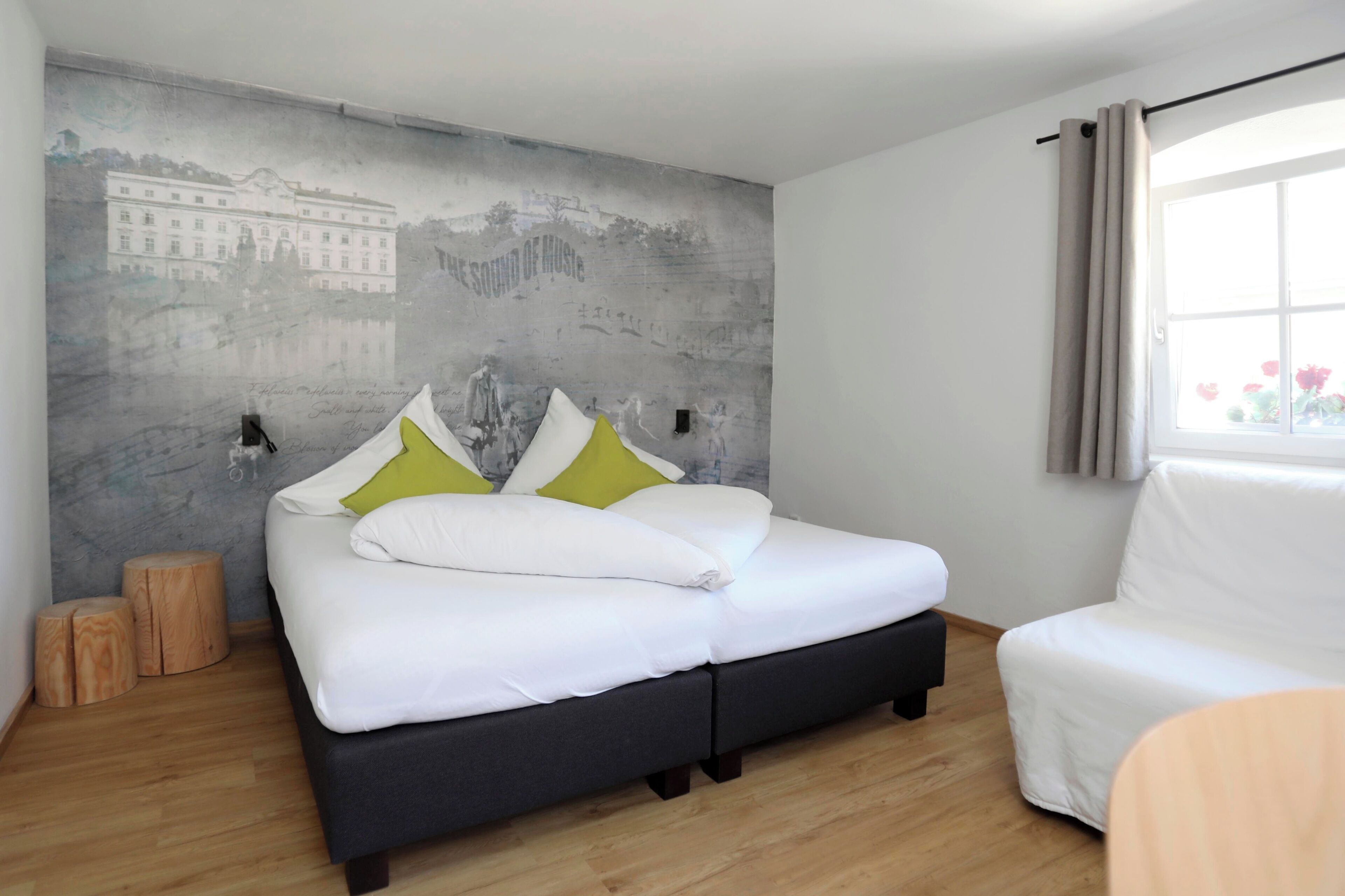 A bedroom with a bed, a chair, a wooden floor, a wall with a mural, and a window with curtains.