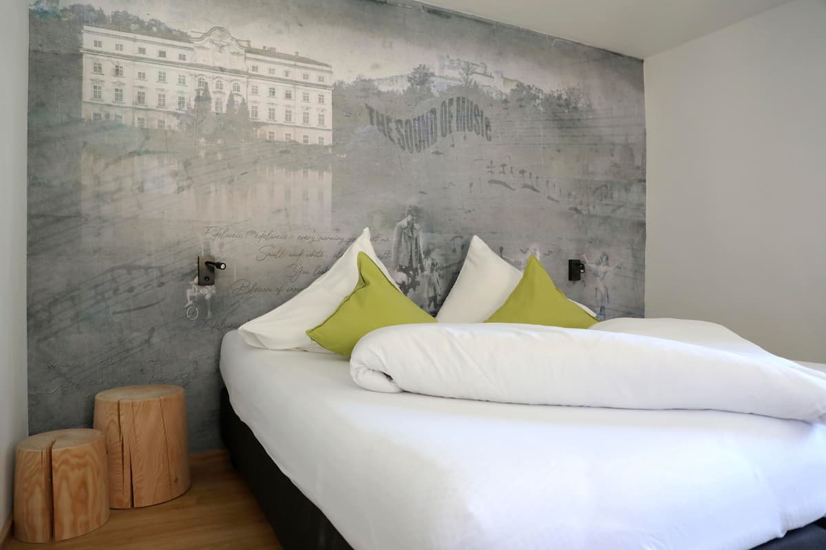 Une chambre avec un grand lit, du linge de lit blanc et deux coussins verts. On y trouve deux tabourets en bois et une fresque murale représentant un bâtiment et un paysage.