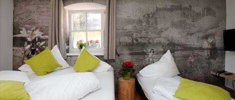 A room with two beds, each with white bedding and green pillows, a small wooden side table with a vase of red flowers, and a window with curtains.