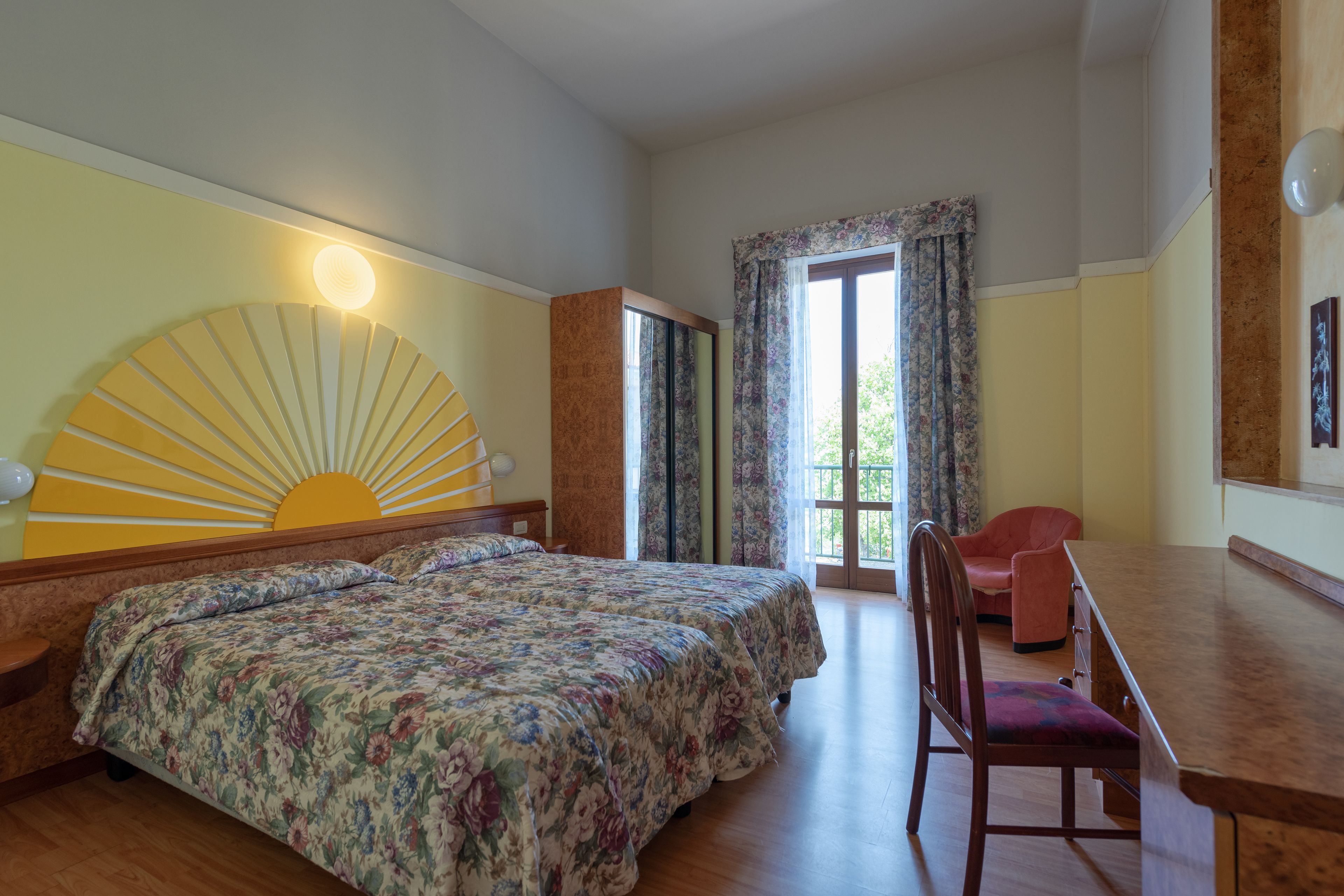 A hotel room with two beds, a desk, and a chair. There is a large window with curtains and a view of greenery outside.