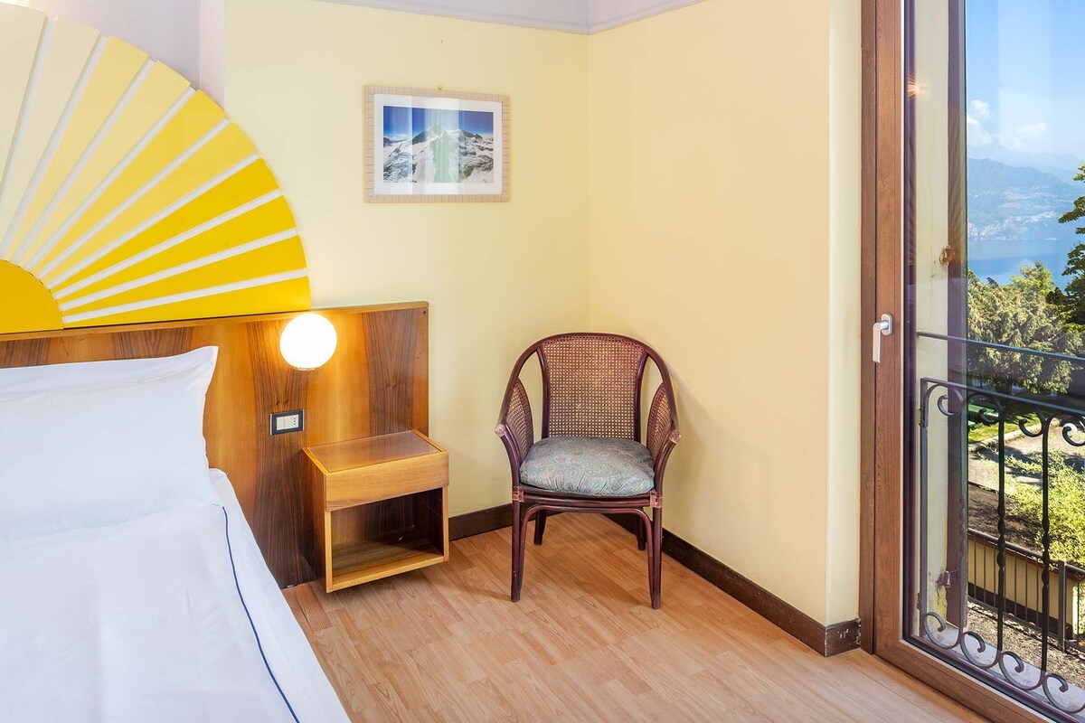 A hotel room with a bed, a chair, a small table, a picture on the wall, and a balcony with a view.