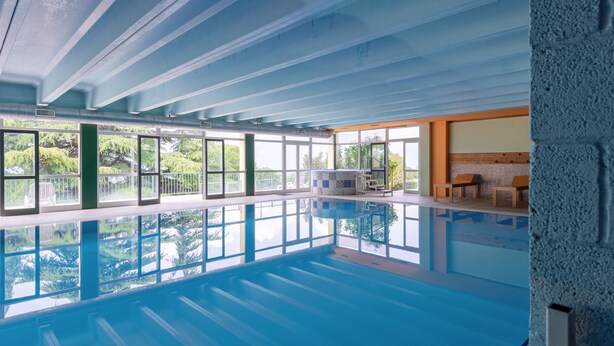 Una piscina interior con paredes de vidrio transparente, suelo de baldosas y una zona de estar con bancos.
