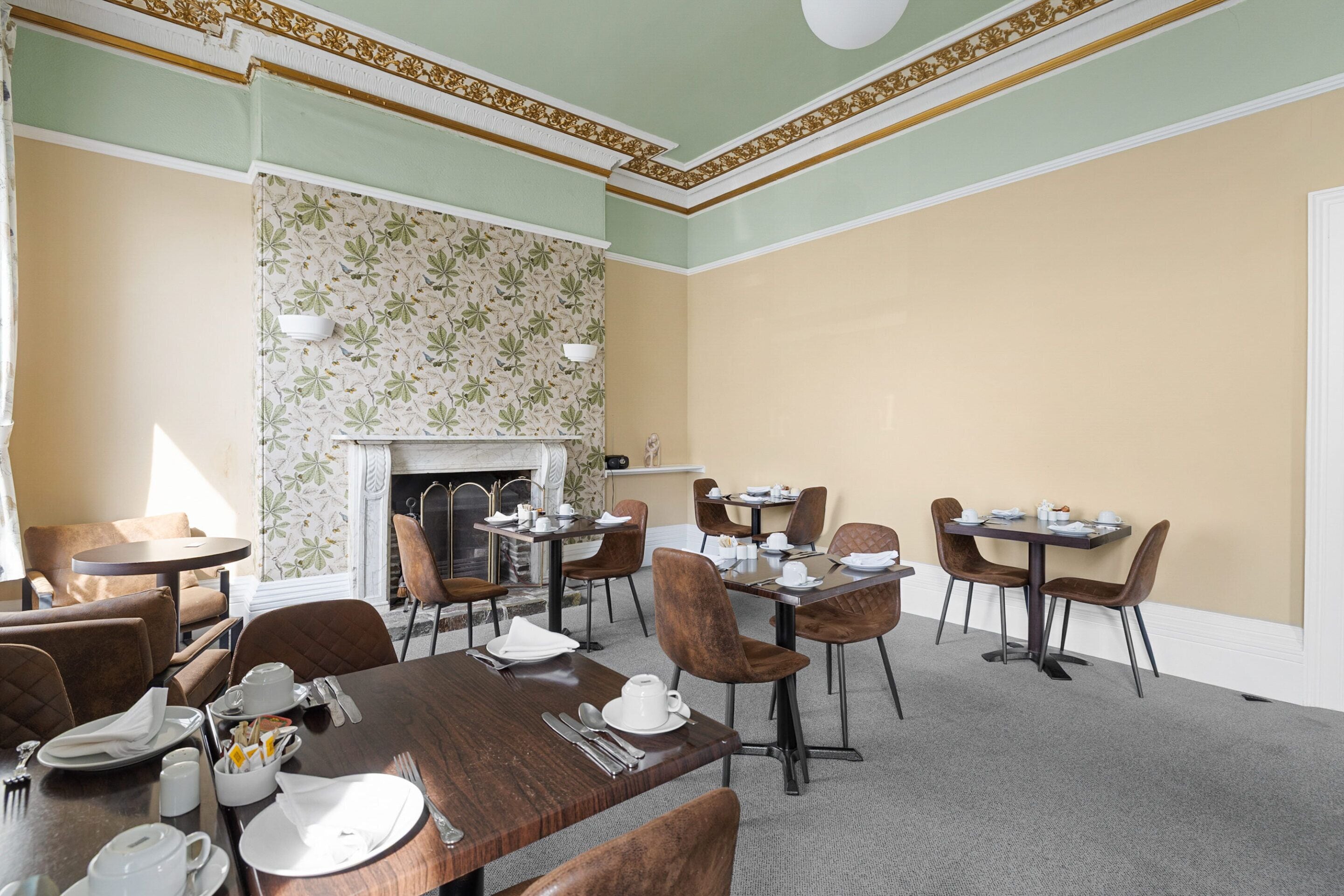 Un espace repas avec des tables et des chaises, une cheminée et un mur recouvert d’un papier peint à motifs.