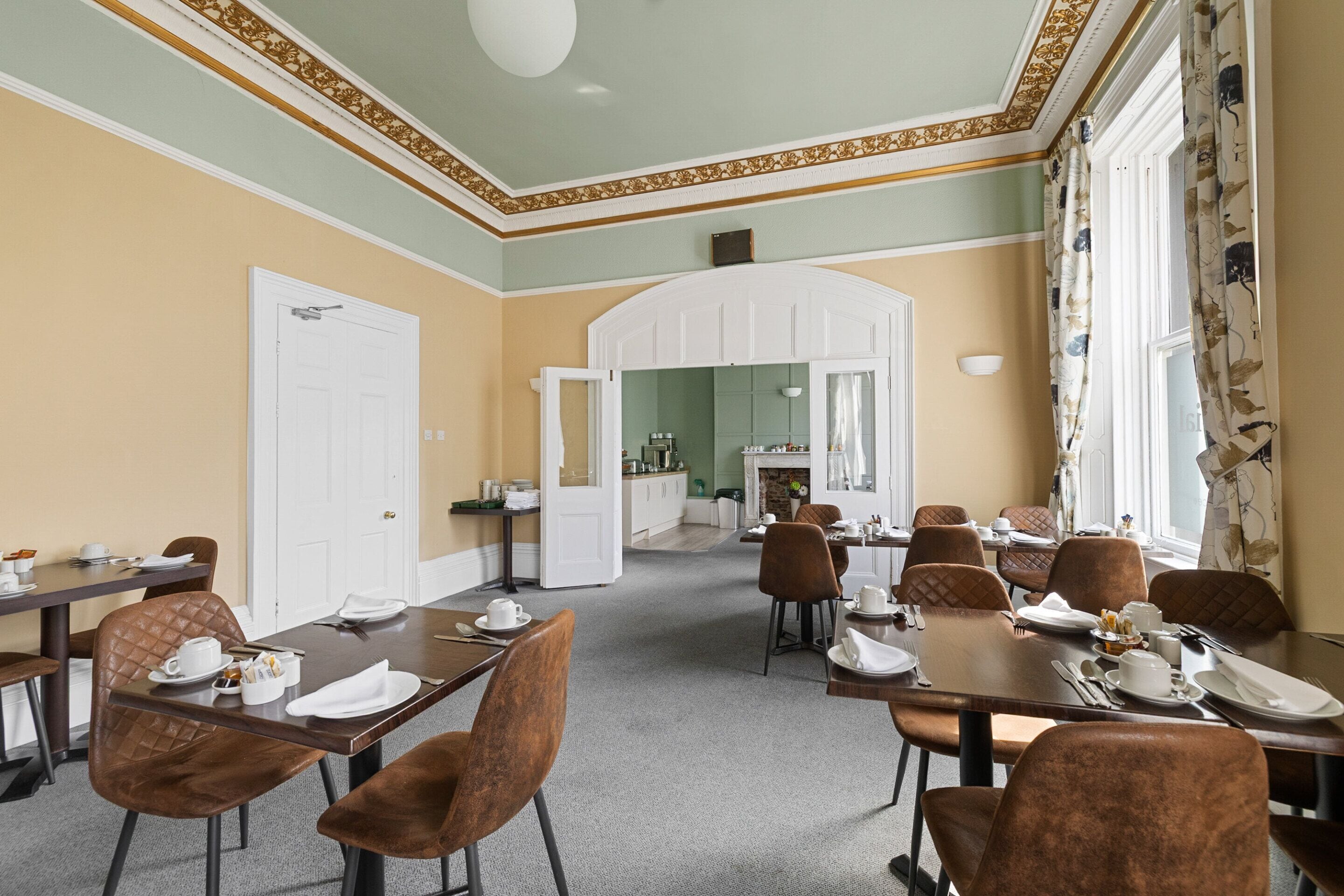 A dining area with tables set for four, brown upholstered chairs, and a view into a kitchen area.