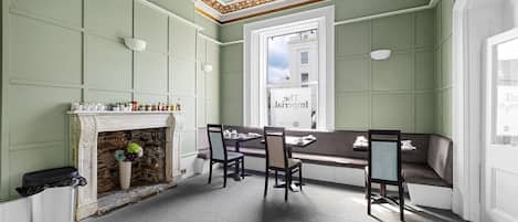 A room with a fireplace, a dining table with chairs, a bench, and a view of the street through a large window.