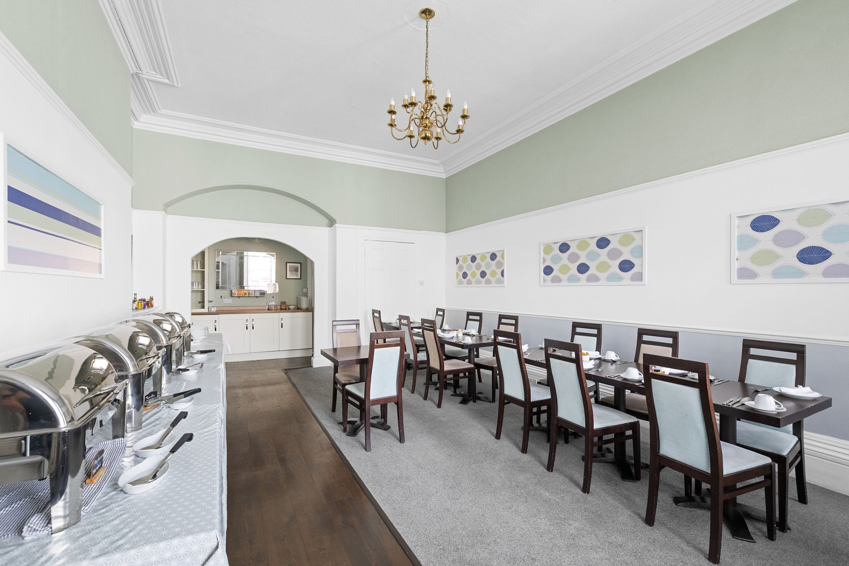 A dining area with multiple tables and chairs, a buffet line with food service equipment, and a chandelier.
