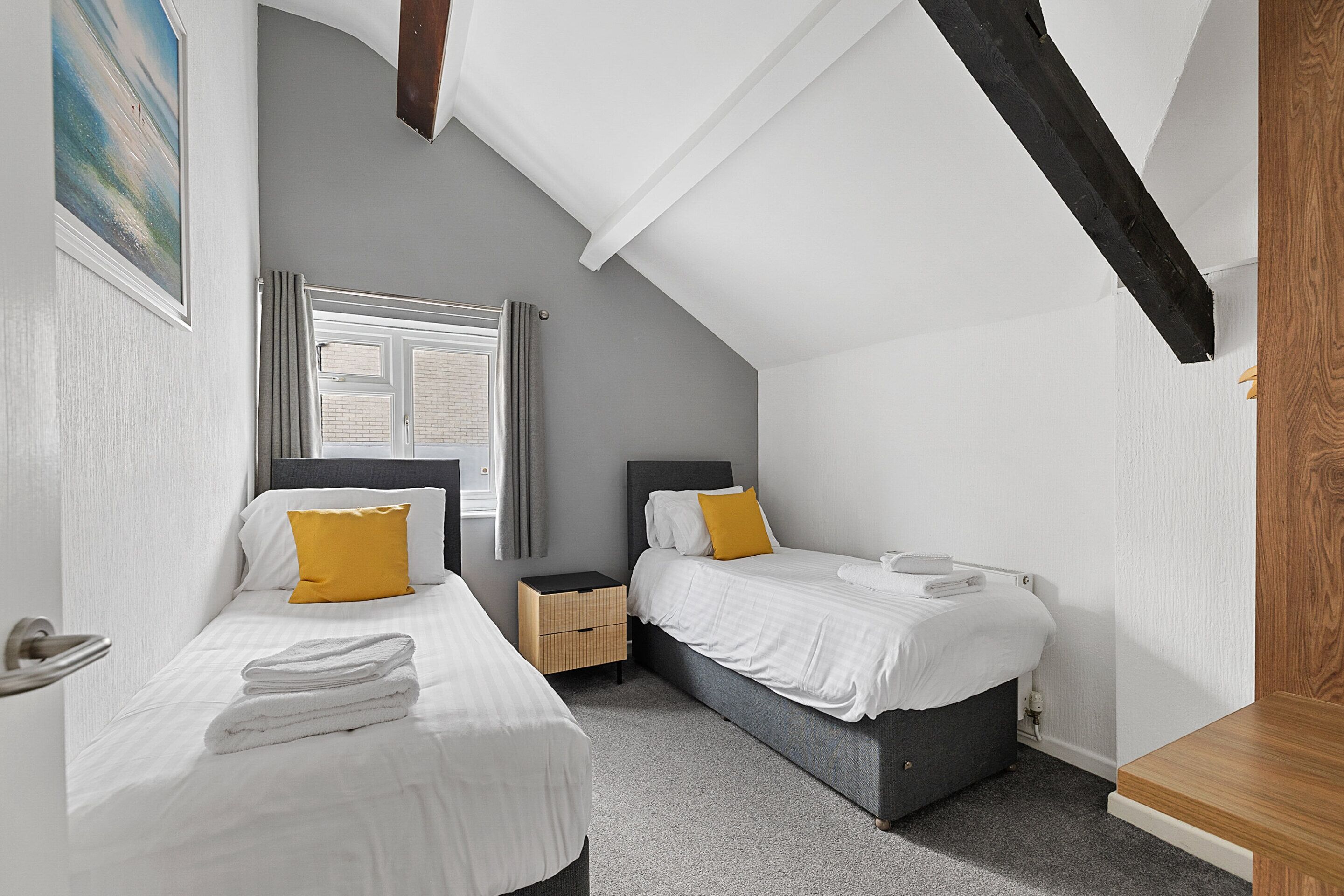 A bedroom with two beds, a window with curtains, a wooden ceiling beam, and a small bedside table.